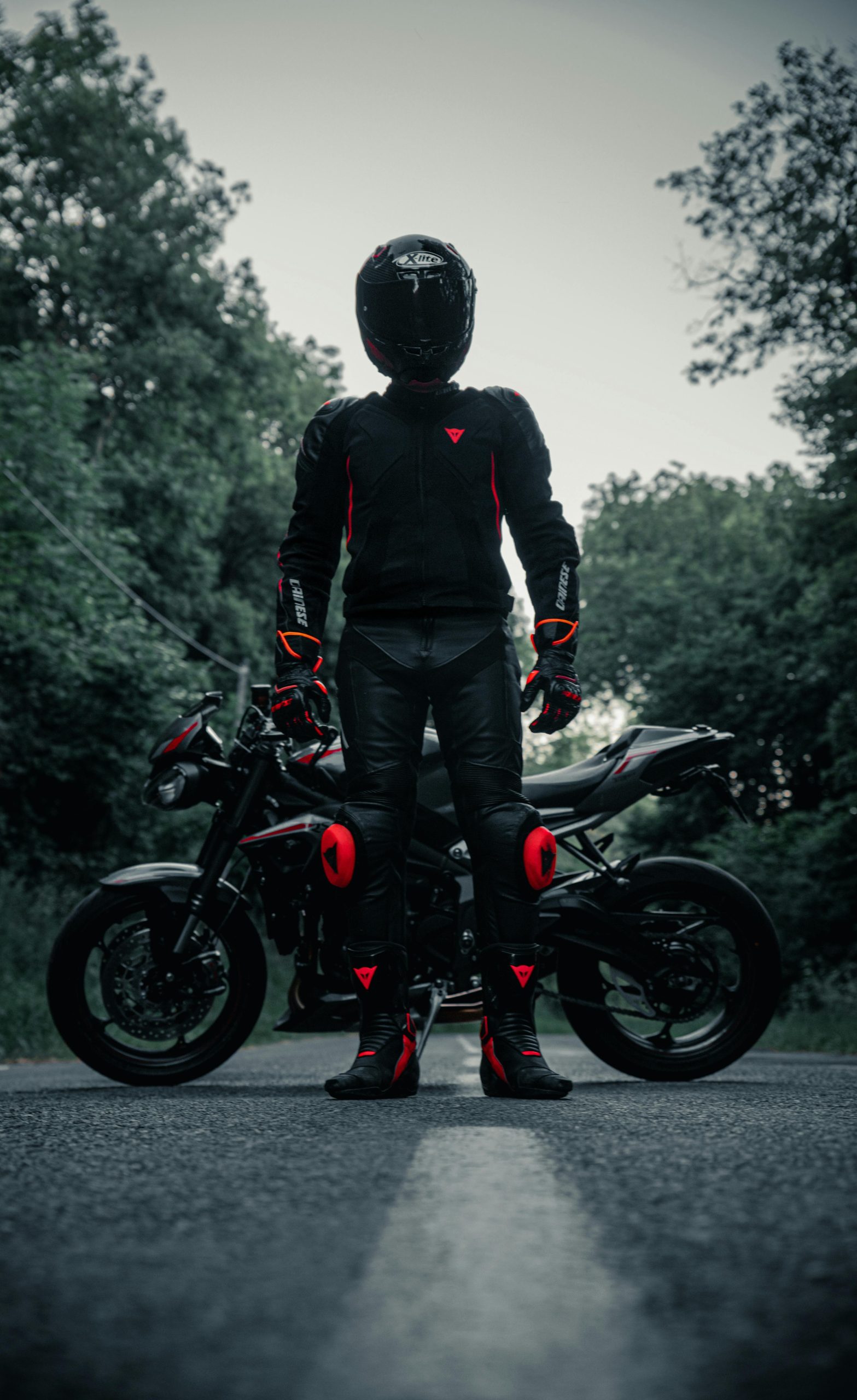 Motorcyclist in black gear and red accents, standing with bike on a French road, outdoors.